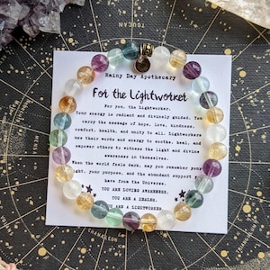May include: A beaded bracelet featuring round, multi-colored translucent stones in shades of purple, yellow, blue, and clear, with a small metallic charm. It rests on a white card printed with the text Rainy Day Apothecary For the Lightworker.