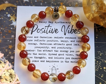 May include: A beaded bracelet with citrine and carnelian stones, alongside a citrine crystal and a card that reads "Positive Vibes." The bracelet features a mix of orange and yellow beads. The card includes text about positivity.