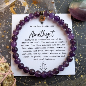 May include: A beaded amethyst bracelet with a small charm is displayed on a white card. The card reads "Rainy Day Apothecary" and "Amethyst." The text describes the stone's healing properties, including stress and anxiety relief.