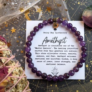 May include: A beaded amethyst bracelet with a gold charm, displayed with a card that reads "Amethyst" and describes its healing properties. A purple and green stone is also visible.