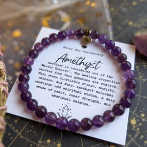 May include: A beaded amethyst bracelet with a silver charm, resting on a card with text. The card reads "Amethyst" and describes the stone's healing properties. A bundle of dried herbs and a purple crystal are also visible.