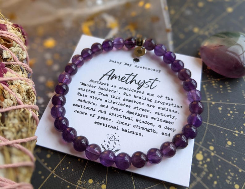 May include: A beaded amethyst bracelet with a silver charm, resting on a card with text. The card reads "Amethyst" and describes the stone's healing properties. A bundle of dried herbs and a purple crystal are also visible.