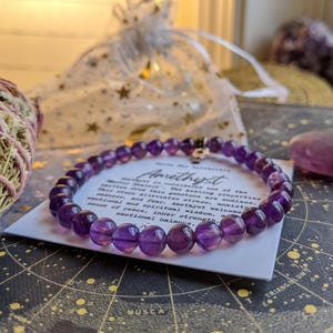 May include: A purple amethyst bead bracelet with a silver charm, resting on a card with text. The card is on a star map, with a small purple crystal and a bundle of sage nearby. A sheer bag with gold stars is in the background.