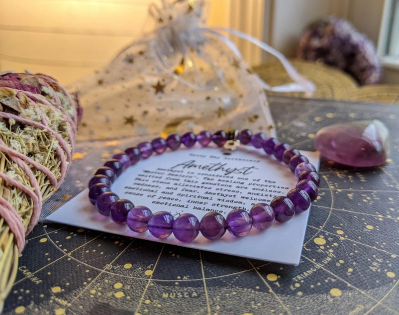 May include: A purple amethyst bead bracelet with a silver charm, resting on a card with text. The card is on a star map, with a small purple crystal and a bundle of sage nearby. A sheer bag with gold stars is in the background.