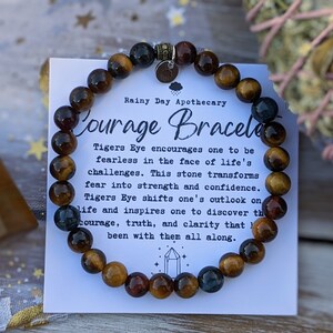 May include: A Courage Bracelet made of tiger's eye beads. The bracelet is displayed with a card that reads "Courage Bracelet" and describes the properties of the stone. The beads are brown, gold, and blue.