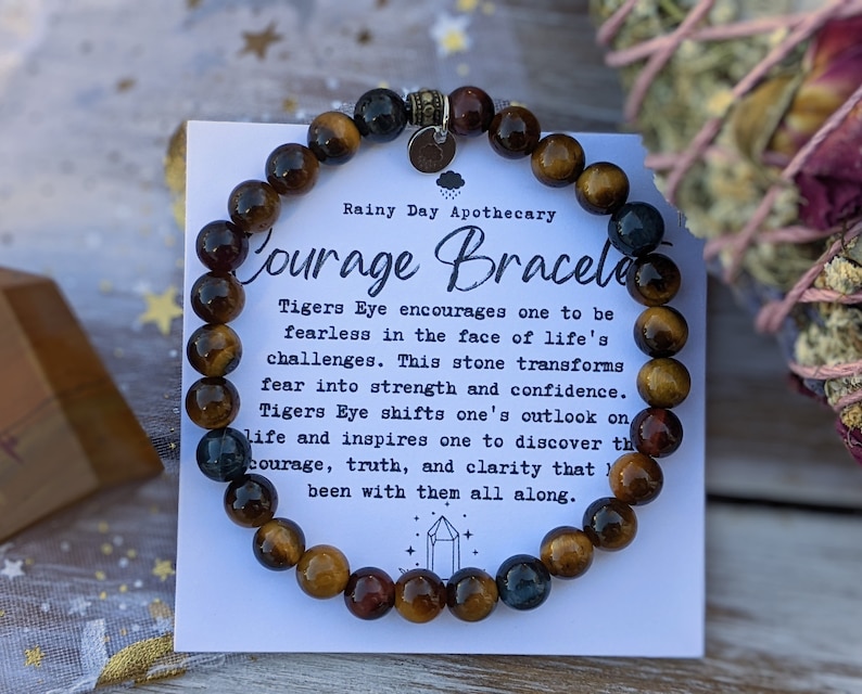 May include: A Courage Bracelet made of tiger's eye beads. The bracelet is displayed with a card that reads "Courage Bracelet" and describes the properties of the stone. The beads are brown, gold, and blue.