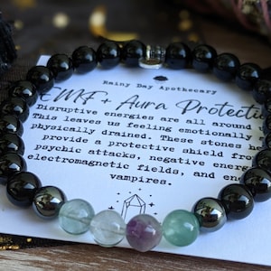 May include: Black onyx, green fluorite, and hematite beads on a bracelet with a silver charm. The bracelet is on a white card with the text "Rainy Day Apothecary EMF+ Aura Protection Disruptive energies are all around This leaves us feeling emotionally physically drained. These stones help provide a protective shield from psychic attacks, negative energies, electromagnetic fields, and energy vampires."