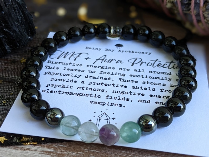 May include: Black onyx, green fluorite, and hematite beads on a bracelet with a silver charm. The bracelet is on a white card with the text "Rainy Day Apothecary EMF+ Aura Protection Disruptive energies are all around This leaves us feeling emotionally physically drained. These stones help provide a protective shield from psychic attacks, negative energies, electromagnetic fields, and energy vampires."