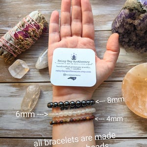 May include: A hand wearing three bracelets with different bead sizes. The top bracelet has 8mm beads, the middle bracelet has 6mm beads, and the bottom bracelet has 4mm beads. The text "all bracelets are made using 6mm beads" is written below the bracelets. The hand is holding a business card that reads "Rainy Day Apothecary" with the text "Inspired to make the world a little brighter." and "handcrafted gemstone jewelry, oils, candles" and "rainydayapothecarync.etsy.com".