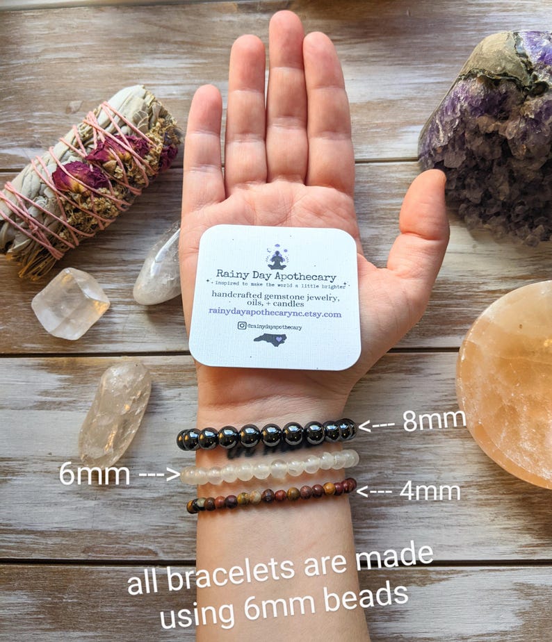 May include: A hand wearing three bracelets with different bead sizes. The top bracelet has 8mm beads, the middle bracelet has 6mm beads, and the bottom bracelet has 4mm beads. The text "all bracelets are made using 6mm beads" is written below the bracelets. The hand is holding a business card that reads "Rainy Day Apothecary" with the text "Inspired to make the world a little brighter." and "handcrafted gemstone jewelry, oils, candles" and "rainydayapothecarync.etsy.com".