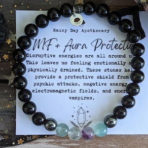 May include: Black beaded bracelet with a charm that says "Rainy Day Apothecary" and the text "SMF + Aura Protection". The text below reads "Disruptive energies are all around us. This leaves us feeling emotionally and physically drained. These stones help provide a protective shield from psychic attacks, negative energy, electromagnetic fields, and energy vampires."