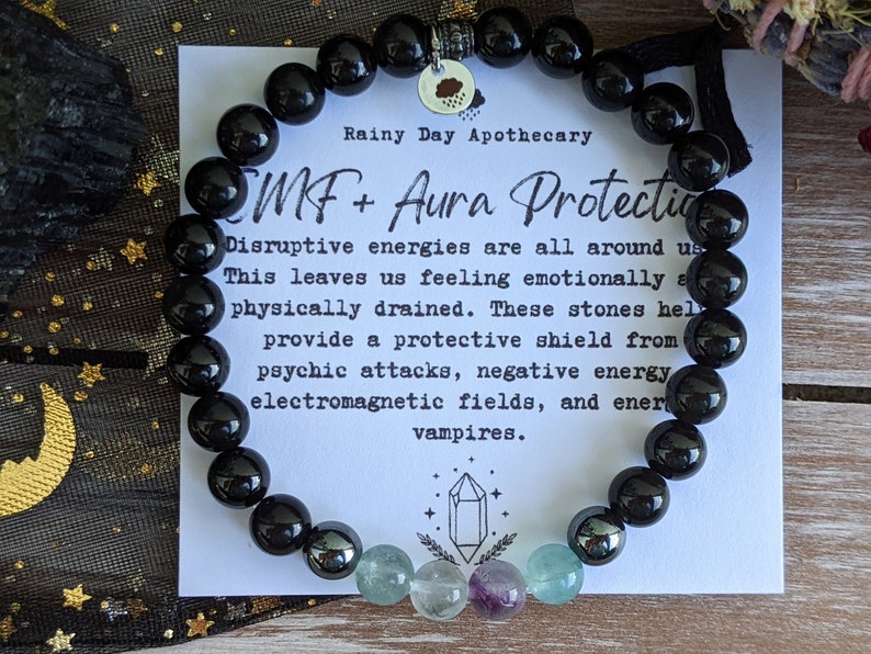 May include: Black beaded bracelet with a charm that says "Rainy Day Apothecary" and the text "SMF + Aura Protection". The text below reads "Disruptive energies are all around us. This leaves us feeling emotionally and physically drained. These stones help provide a protective shield from psychic attacks, negative energy, electromagnetic fields, and energy vampires."