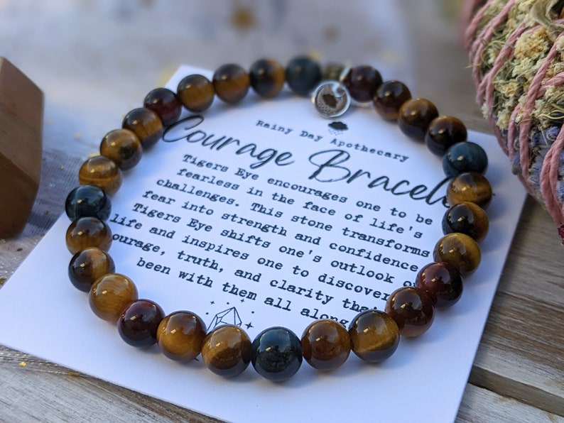 May include: A Courage Bracelet made of tiger's eye stones. The bracelet is displayed on a card with the text "Courage Bracelet" and a description of the stone's properties. The bracelet is a mix of brown, gold, and dark blue beads.