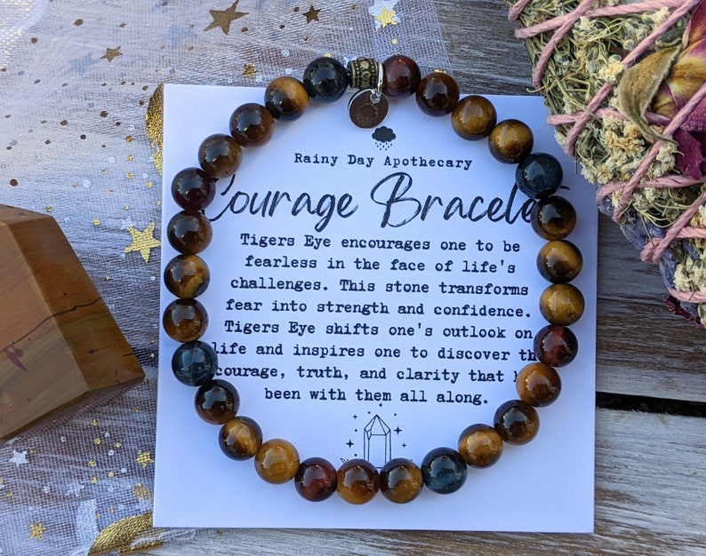 May include: A Courage Bracelet made of tiger's eye beads. The bracelet is displayed with a card that reads "Courage Bracelet" and describes the properties of tiger's eye. The beads are a mix of brown, gold, and blue.