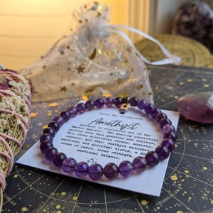 May include: A beaded amethyst bracelet with a small gold charm, displayed on a card with text about the stone's properties. The image includes a bundle of sage, a small amethyst stone, and a sheer bag with star-shaped accents.