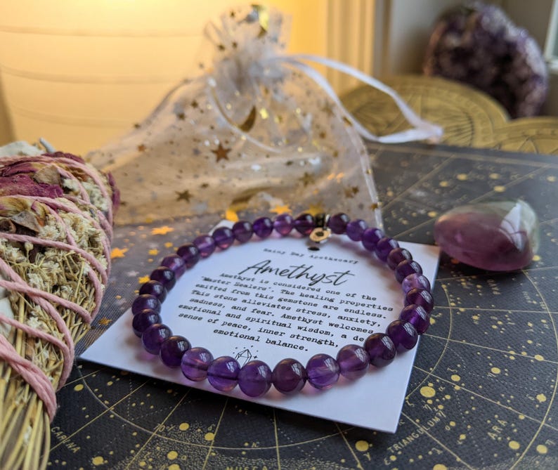 May include: A beaded amethyst bracelet with a small gold charm, displayed on a card with text about the stone's properties. The image includes a bundle of sage, a small amethyst stone, and a sheer bag with star-shaped accents.