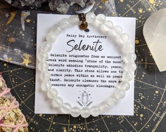 May include: A selenite beaded bracelet with a small charm is displayed on a card with the text "Selenite." The card describes the stone's properties. The bracelet is made of translucent, round beads.