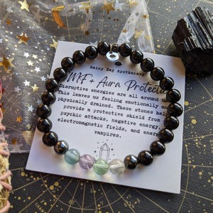 May include: Black onyx beaded bracelet with a charm and a card that reads "MF + Aura Protection". The card explains that the bracelet helps protect against disruptive energies, psychic attacks, negative energy, electromagnetic fields, and energy vampires.