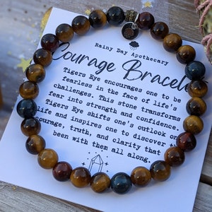 May include: A Courage Bracelet made of tiger's eye beads in shades of brown, gold, and blue. The bracelet is displayed with a card that reads "Courage Bracelet" and describes the properties of tiger's eye.
