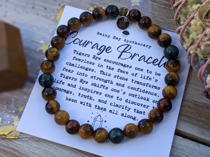May include: A Courage Bracelet made of tiger's eye beads in shades of brown, gold, and blue. The bracelet is displayed with a card that reads "Courage Bracelet" and describes the properties of tiger's eye.
