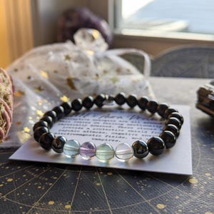 May include: Black onyx and rainbow fluorite beaded bracelet with a silver clasp. The bracelet is on a white card with text that reads "All that protects all around us, bringing us peace and tranquility. We listen to our feelings, we emotionally protect ourselves. These shield from psychic attacks, negative energies, electromagnetic fields, and energy vampires."