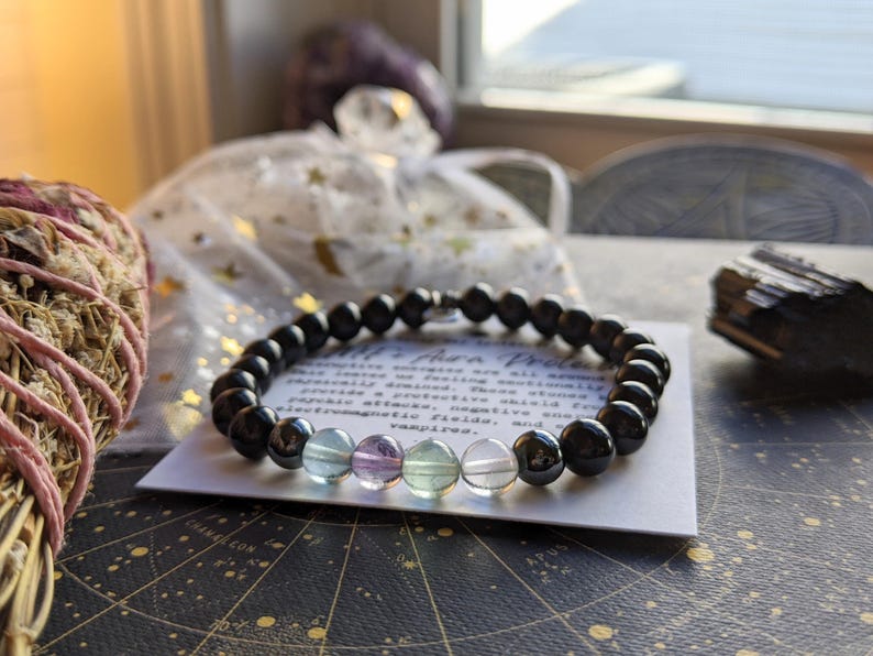May include: Black onyx and rainbow fluorite beaded bracelet with a silver clasp. The bracelet is on a white card with text that reads "All that protects all around us, bringing us peace and tranquility. We listen to our feelings, we emotionally protect ourselves. These shield from psychic attacks, negative energies, electromagnetic fields, and energy vampires."
