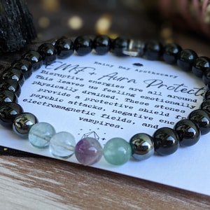 May include: Black beaded bracelet with green, purple, and clear stones. The bracelet is on a white card with the text "CMF+ Aura Protection" and "Rainy Day Apothecary". Disruptive energies are all around. This leaves us feeling emotionally and physically drained. These stones provide a protective shield from psychic attacks, negative electromagnetic fields, and vampires.