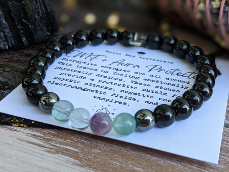 May include: Black beaded bracelet with green, purple, and clear stones. The bracelet is on a white card with the text "CMF+ Aura Protection" and "Rainy Day Apothecary". Disruptive energies are all around. This leaves us feeling emotionally and physically drained. These stones provide a protective shield from psychic attacks, negative electromagnetic fields, and vampires.
