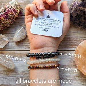 May include: Three gemstone bracelets are displayed on a wrist, with bead sizes of 4mm, 6mm, and 8mm. The bracelets are made with 6mm beads. The image includes a card from Rainy Day Apothecary, offering handcrafted gemstone jewelry, oils, and candles.