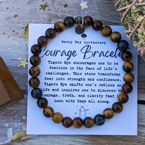 May include: A Courage Bracelet made of Tiger's Eye beads. The bracelet is displayed with a card that reads "Courage Bracelet" and describes the stone's properties. The beads are a mix of brown, gold, and blue hues. A wooden crystal and sage bundle are also in the image.