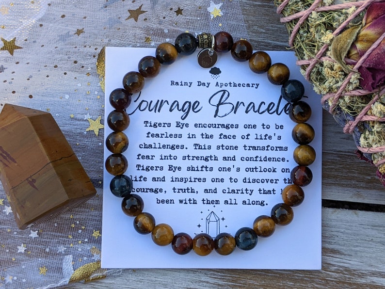 May include: A Courage Bracelet made of Tiger's Eye beads. The bracelet is displayed with a card that reads "Courage Bracelet" and describes the stone's properties. The beads are a mix of brown, gold, and blue hues. A wooden crystal and sage bundle are also in the image.