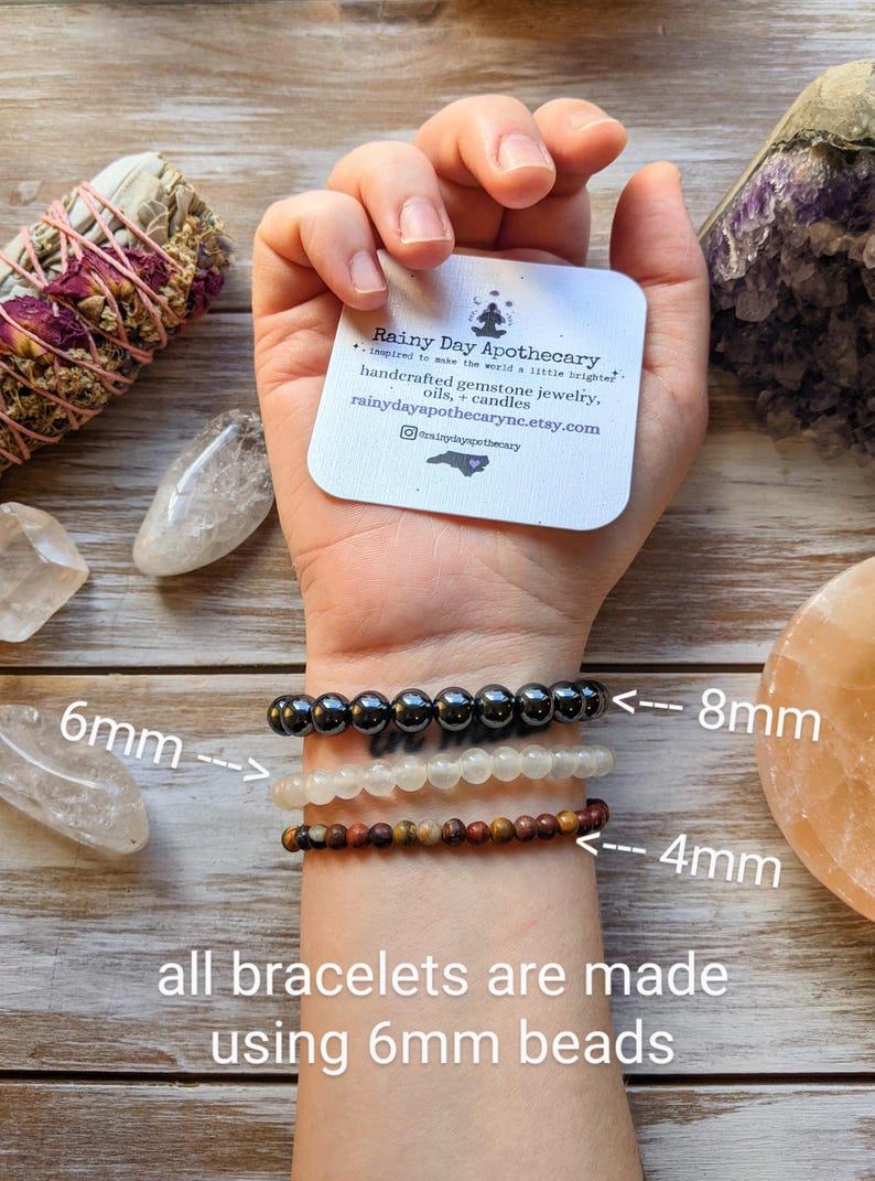 May include: Three gemstone bracelets are displayed on a wrist, with bead sizes of 4mm, 6mm, and 8mm. The bracelets are made with 6mm beads. The image includes a card from Rainy Day Apothecary, offering handcrafted gemstone jewelry, oils, and candles.