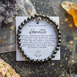 May include: A hematite bracelet with dark gray beads is displayed on a star chart. A card next to the bracelet reads "Rainy Day Apothecary Hematite" with a description of the gemstone. Crystals and other items surround the bracelet.