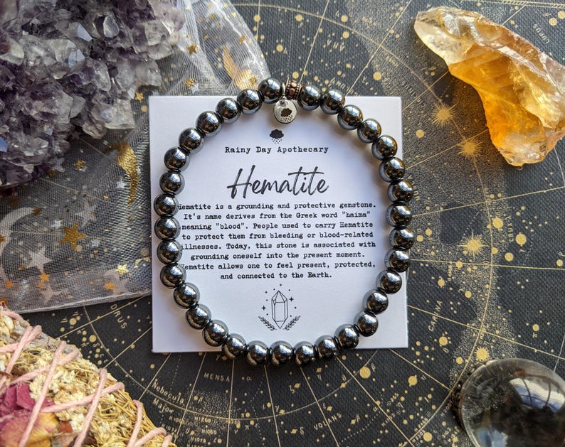 May include: A hematite bracelet with dark gray beads is displayed on a star chart. A card next to the bracelet reads "Rainy Day Apothecary Hematite" with a description of the gemstone. Crystals and other items surround the bracelet.