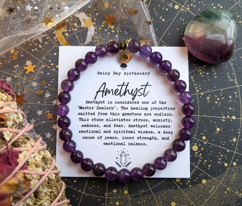 May include: A beaded amethyst bracelet with a gold charm, displayed with a card that reads "Amethyst" and describes its healing properties. A purple and green stone is also visible.