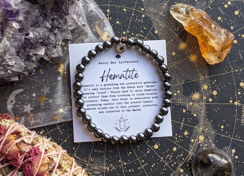 May include: A hematite bead bracelet with a small charm, displayed with a card from Rainy Day Apothecary. The card describes hematite as a grounding gemstone. Other crystals and a bundle of dried flowers are also in the image.