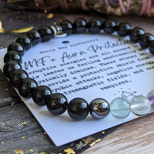 May include: Black beaded bracelet with a silver charm and a card that reads "MF+ Aura Protection" with text about the protective properties of the stones. The bracelet has black beads, a hematite bead, and green and purple fluorite beads.