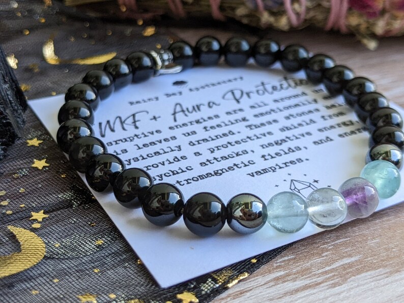 May include: Black beaded bracelet with a silver charm and a card that reads "MF+ Aura Protection" with text about the protective properties of the stones. The bracelet has black beads, a hematite bead, and green and purple fluorite beads.