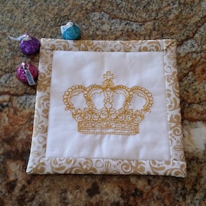 May include: A white square coaster with a gold embroidered crown design. The coaster has a gold patterned border.