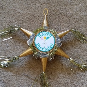 May include: A gold star-shaped pinata with a turquoise and gold circle that says "Happy Birthday" with three balloons in the center. The pinata has gold fringe and gold tassels.