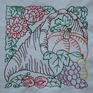 May include: Embroidered design featuring a cornucopia overflowing with autumn harvest symbols. A large pumpkin, grapes, and a pear are surrounded by leaves and flowers in shades of red, green, brown, and orange.