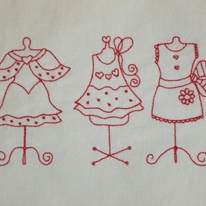 May include: Three red embroidered dresses on display stands. Each dress has a different style and features small details like hearts and polka dots.