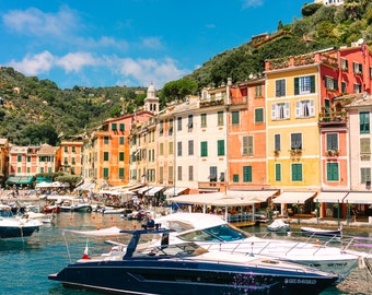 May include: A picturesque harbor scene with colorful buildings lining the waterfront. Several boats are docked in the water, including a large white yacht and a smaller black speedboat.
