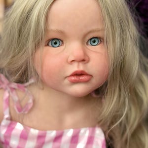 May include: A lifelike doll with fair skin, blue eyes, and long, wavy blonde hair. The doll is wearing a pink and white checkered dress with ribbon straps. The doll's face has a realistic appearance with detailed features.