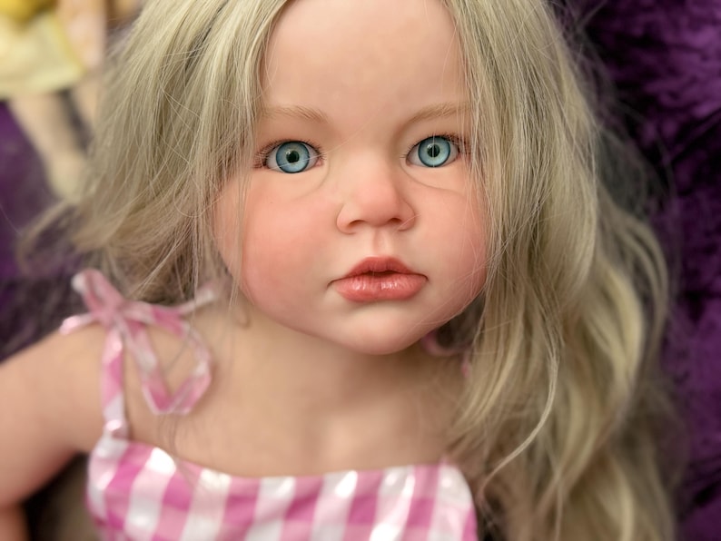 May include: A lifelike doll with fair skin, blue eyes, and long, wavy blonde hair. The doll is wearing a pink and white checkered dress with ribbon straps. The doll's face has a realistic appearance with detailed features.