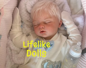 May include: A realistic baby doll with closed eyes, wearing a white onesie with a blue bow and a decorative crown. The doll has light blonde hair and is lying in a soft, white blanket. A bottle is next to the doll. The text "Lifelike Dolls" is in yellow.