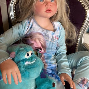 May include: A young girl with blonde hair and blue eyes wearing a light blue pajama set featuring Elsa from Frozen. She is holding a large, teal Sully plush toy from Monsters, Inc. The plush has purple spots and brown claws.