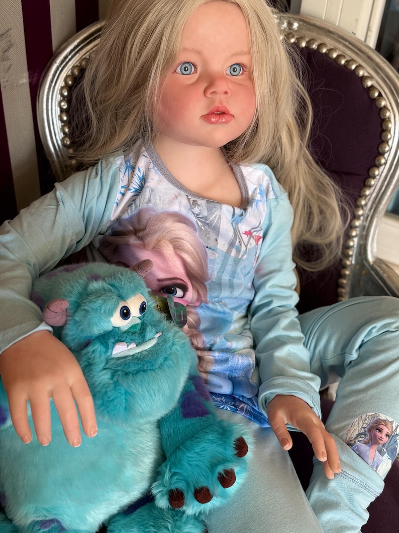 May include: A young girl with blonde hair and blue eyes wearing a light blue pajama set featuring Elsa from Frozen. She is holding a large, teal Sully plush toy from Monsters, Inc. The plush has purple spots and brown claws.