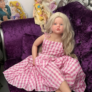 May include: A lifelike doll with long blonde hair and blue eyes, wearing a pink and white gingham dress. The doll is seated on a plush purple velvet couch. Additional dolls and character cutouts are visible in the background.