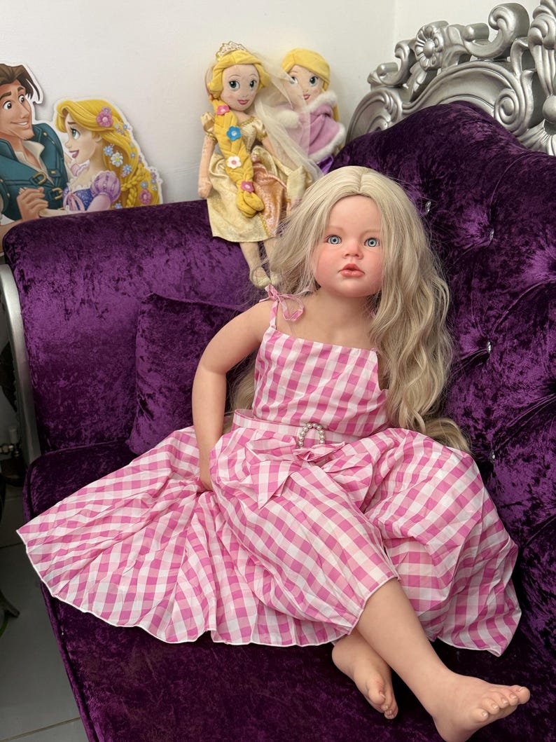 May include: A lifelike doll with long blonde hair and blue eyes, wearing a pink and white gingham dress. The doll is seated on a plush purple velvet couch. Additional dolls and character cutouts are visible in the background.
